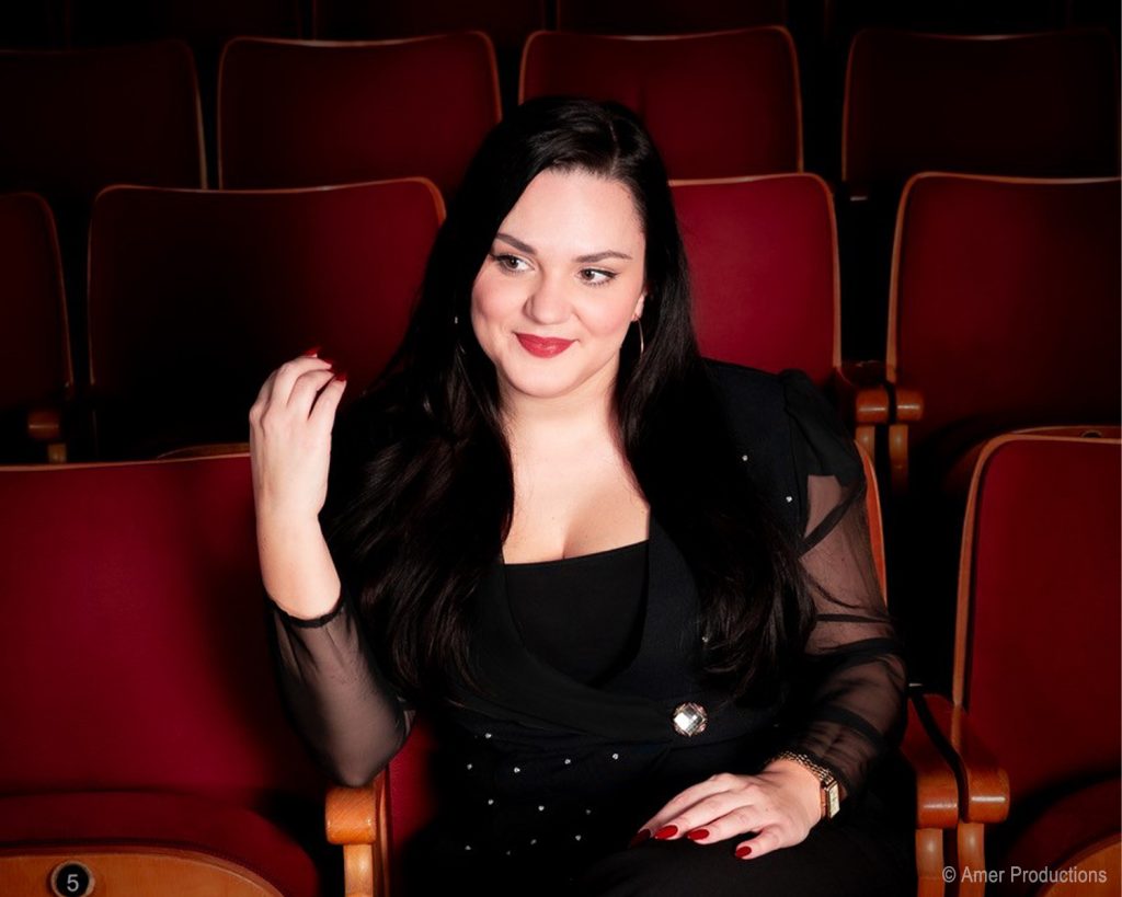 Ina Jovanovic sitzt alleine in einem roten Theatersessel, trägt schwarze Kleidung und lächelt nach links.