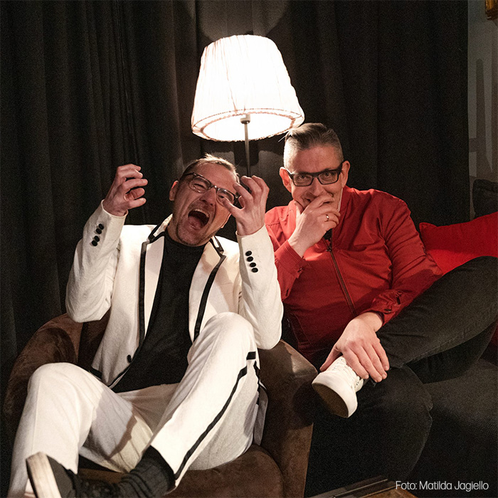 Gebrüder Moped auf einem Sofa vor schwarzem Vorhang und einer großen Stehlampe. Martin Moped links trägt einen weißen Anzug mit schwarzem Shirt und reißt mit weit geöffnetem Mund und erhobenen Händen theatralisch die Augen auf. Franz Moped rechts trägt eine rote Jacke, sitzt entspannt daneben und hält die Hand nachdenklich vor den Mund.